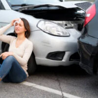 Woman sitting on the ground leaning against her damaged car holding her head. Indicating a head injury