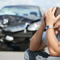 A man sitting with his hands in his head and a crashed black car in the background